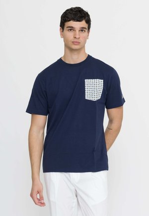 Jonge man met donker krullend haar, die een marineblauw T-shirt draagt met een wit patroon op de zak en een witte broek, staand tegen een effen achtergrond.