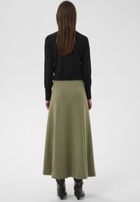 Falda midi verde de textura suave, cintura alta y diseño en línea A, combinada con un top negro ajustado de manga larga y botas negras hasta la rodilla.