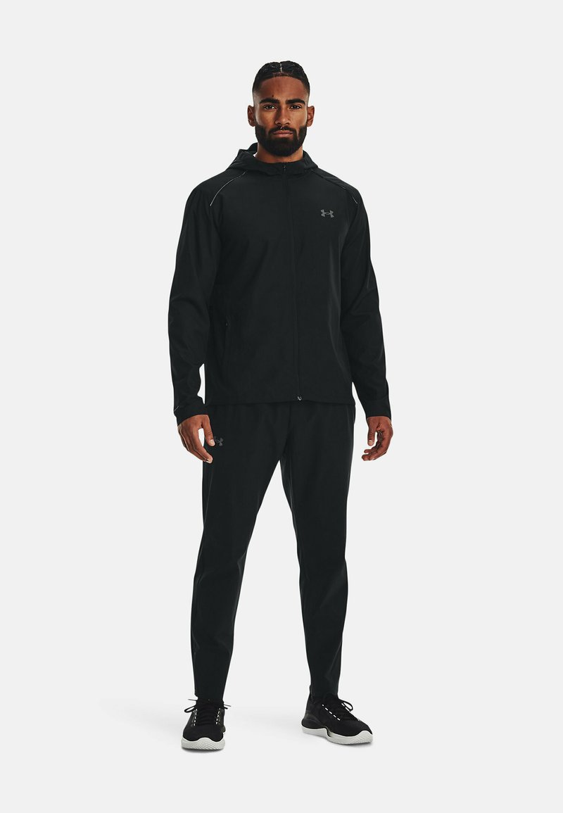 Veste de survêtement noire zippée avec capuche, pantalons noirs assortis et chaussures noires. Le matériau semble lisse avec des accents minimes et un design épuré.