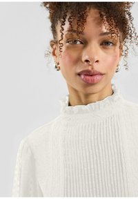 Witte getextureerde blouse met gerimpelde kraag, voorzien van verticale plooien en kleine verhoogde stippen. Model draagt zilveren oorringen.