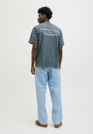 Mann mit kurzem lockigem Haar, der ein dunkelgraues T-Shirt mit Grafik, hellblaue Jeans und braune Schuhe trägt, steht mit dem Rücken zur Kamera.