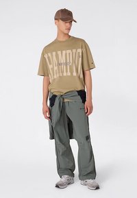 T-shirt kaki avec le mot "CHAMPION" en grosses lettres crème. Pantalon vert olive, coupe décontractée avec poches. Baskets grises et casquette marron.