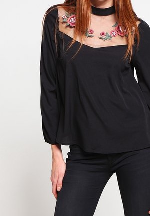 Nainen pukeutunut mustaan pitkähihaseen yläosaan, jossa on läpinäkyvä kaulus ja vaaleanpunainen kukkaistikkaus, yhdistettynä mustiin housuihin.