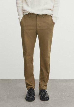 Homme portant un pantalon droit marron, un pull blanc côtelé et des chaussures à lacets noires, debout les mains dans les poches sur un sol gris.