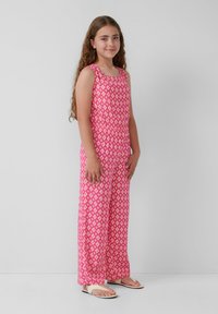 Rosa ärmelloser Jumpsuit mit einem Diamantmuster, elastischem Bund und weit geschnittenem Bein. Kombiniert mit leichten Sandalen.