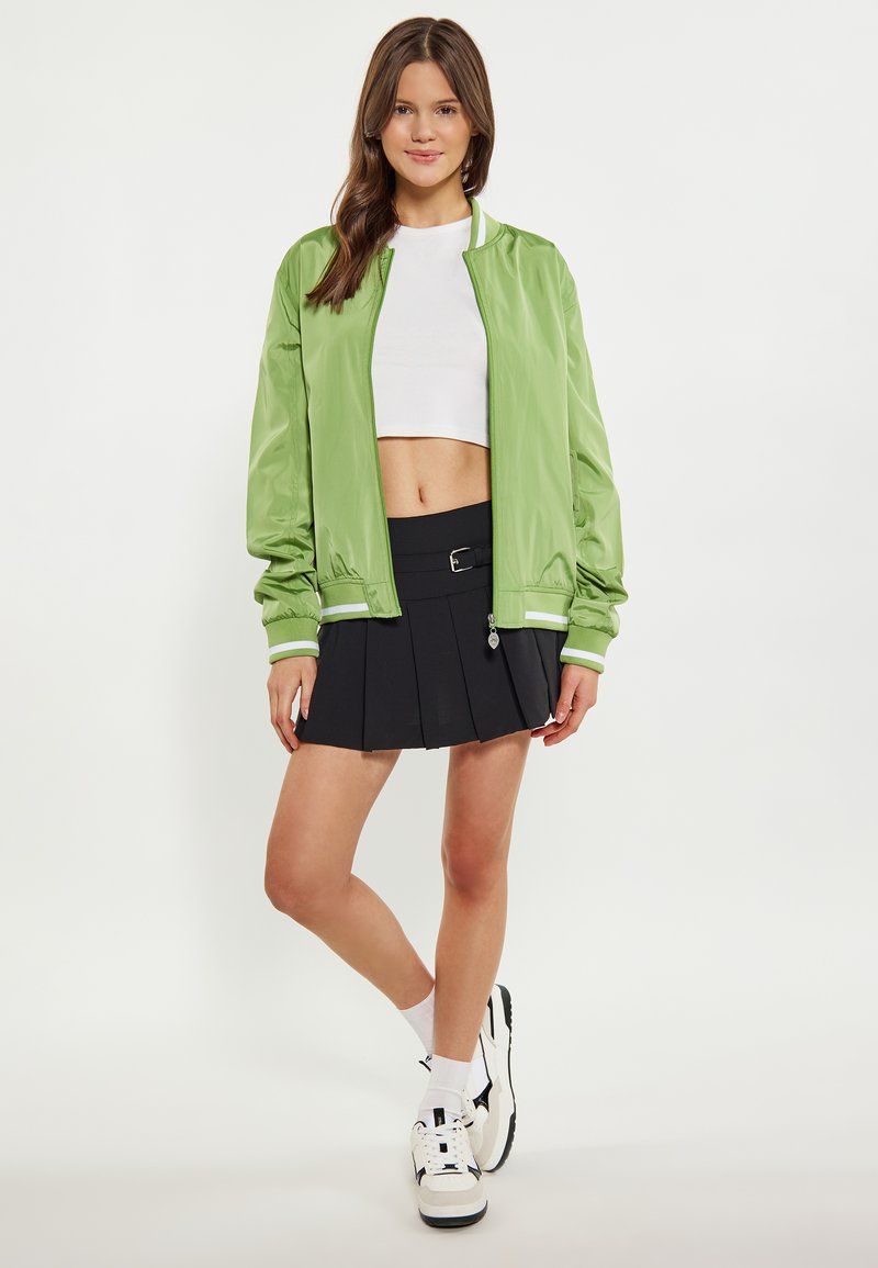 Giacca bomber verde chiaro con polsini bianchi a contrasto, abbinata a una gonna plissettata nera e sneaker bianche. Texture liscia, design atletico.