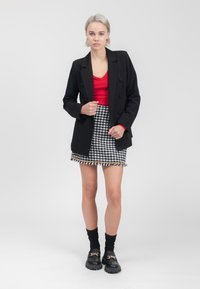 Schwarze maßgeschneiderte Blazer mit doppelten Knöpfen, rotem V-Ausschnitt-Oberteil, Rock mit Hahnentritt-Muster und Fransen, schwarze klobige Loafers und gerippte Socken.