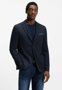 Granatowy, teksturowany blezer z dwoma przednimi kieszeniami, zapięciem na jeden guzik i ozdobną poszetką. Połączony z ciemnoniebieskimi dżinsami.