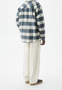 Camicia di flanella a quadri blu e crema, con colletto e polsini con bottone. Abbinata a pantaloni bianchi a gamba larga e scarpe nere.