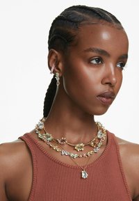 Ženska z zapletenimi lasmi nosi večplastno, barvito nakita iz dragih kamnov in textured rjavordečo bluzo, kar prikazuje samozavesten izraz.