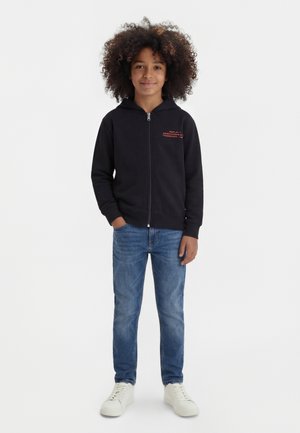 Lapsi, jolla on kiharat hiukset ja musta vetoketjullinen huppari, siniset farkut ja valkoiset lenkkitossut, seisoo yksiväristä valkoista taustaa vasten.