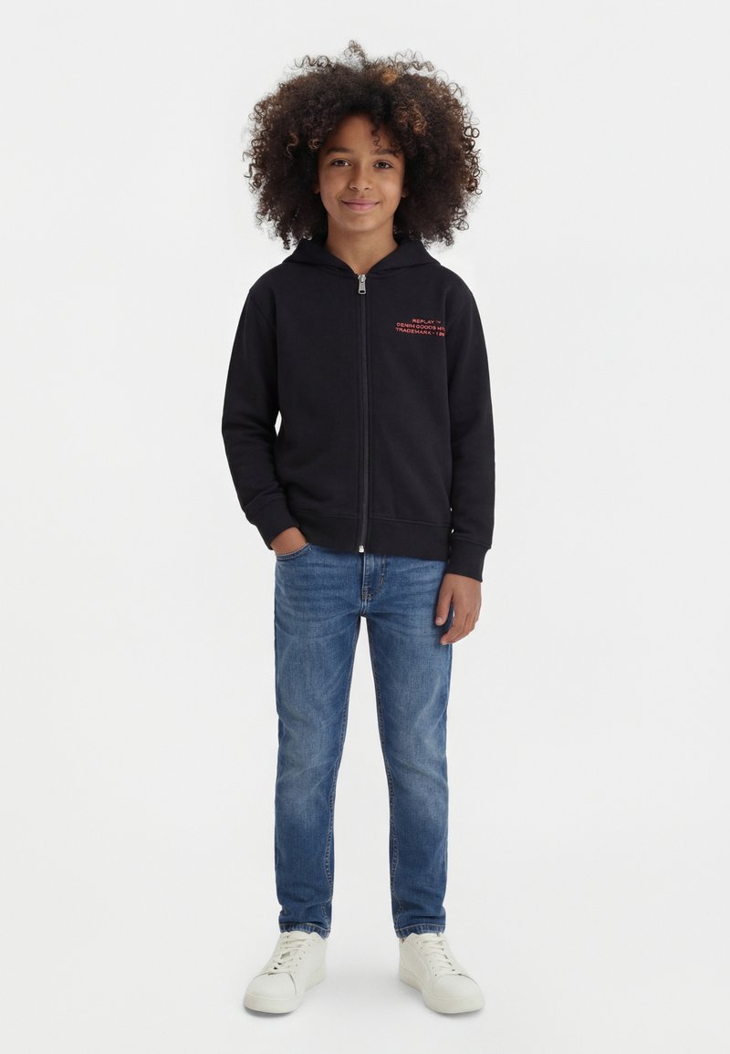 Παιδί με σγουρά μαλλιά που φοράει μαύρο φούτερ με φερμουάρ, μπλε τζιν και λευκά αθλητικά παπούτσια, στέκεται μπροστά σε ένα απλό λευκό φόντο.