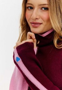 Młoda kobieta ubrana w burgundowy i różowy sweter z niebieskim sercem na rękawie, delikatnie się uśmiechająca, z luźnymi blond włosami.