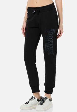 Unterkörper trägt schwarze Joggerhosen mit dem Schriftzug „Boxeur Des Rues“ am Bein und weiße Sneakers mit schwarzen Schnürsenkeln und Fersendetails.