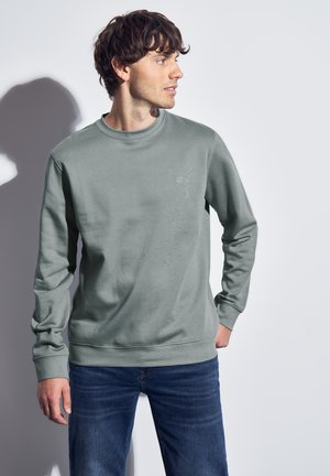 Mand i en salviegrøn sweatshirt og blå jeans, der kigger til højre, stående mod en ensfarvet lys grå baggrund.