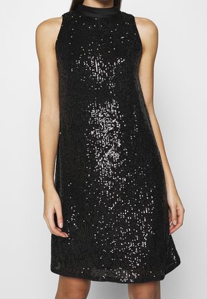 Femme portant une robe noire sans manches avec un col montant et des sequins noirs brillants éparpillés sur le tissu.