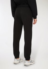 Osoba ubrana w czarne spodnie dresowe i srebrne buty do biegania, stojąca na jednolitym białym tle.