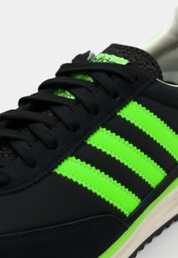Black athletic shoe with green three-stripe design. Features textured fabric, flat laces, and rubber sole with serrated edges.