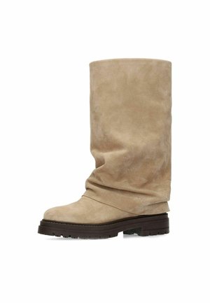 Beige suède midkuitlaars met losse schacht, afgeronde neus en dikke donkerbruine rubberen profielzool.