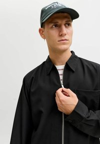 Giacca nera con zip, collo e tasca frontale, indossata sopra una maglietta a righe. Cappellino verde scuro con scritte bianche.