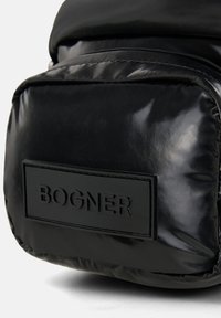Sort polstret nylon taske med en glansfuld overflade, der har et hævet rektangulært label med "BOGNER" præget i fede bogstaver.