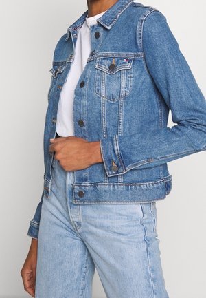 Person trägt eine hellblaue Jeansjacke über einem weißen Hemd und dazu passende hellblaue Jeans, Hand hält die Jacke in der Nähe der Taille.