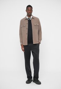 Beige stoffjakke med to brystlommer, lagd over en svart genser og parret med svarte bukser og sko. Nøytral fargepalett.