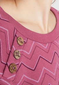 Primer plano de una camisa de patrón zigzag púrpura con tres botones de madera, uno grabado con una hoja, que se lleva en el cuello y la parte superior del pecho de una persona.