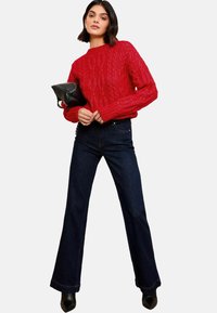 Piros, kötött pulóver hosszú ujjakkal, sötétkék, bővebb szárú farmernadrággal párosítva, fekete clutch táskával és fekete, hegyes orrú magassarkúval kiegészítve.