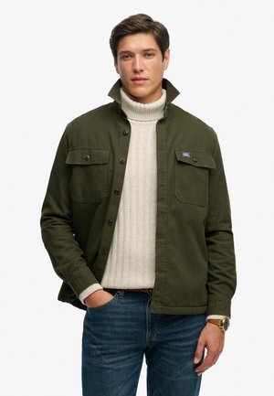Ung mand med mørkt hår iført en olivengrøn knappet jakke over en hvid, kabelstrikket rullekrave og blå jeans.