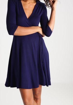 Kobieta ubrana w granatową sukienkę kopertową sięgającą kolan z rękawami do łokcia, stoi z jedną ręką skrzyżowaną, a drugą dotyka włosów.