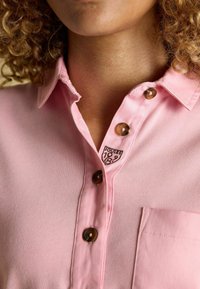 Heled roosa nupuga särk, mis on valmistatud tekstuuritud kangast, hõbedaste nööpidega, tasku ja väike must logo krae lähedal.