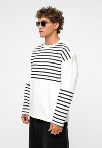 Witte longsleeve shirt met zwarte horizontale strepen over de borst en mouwen. Zacht materiaal, relaxte pasvorm, ronde hals.