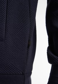 Granatowy sweter dzianinowy z diagonalnym wzorem teksturowym, ściągaczami przy mankietach i małą czarną metką z logo. Miękka tkanina o dopasowanym kroju.
