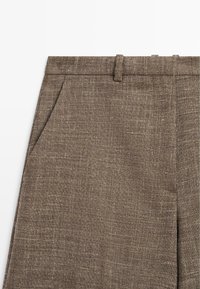 Gros plan sur un pantalon en tissu marron texturé avec passants de ceinture et poches latérales.