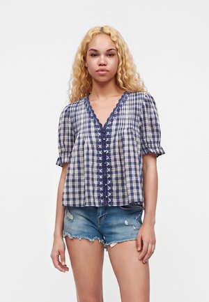 Jeune femme aux longs cheveux blonds bouclés portant un chemisier à carreaux bleu et blanc avec bordure en dentelle et un short en jean usé.