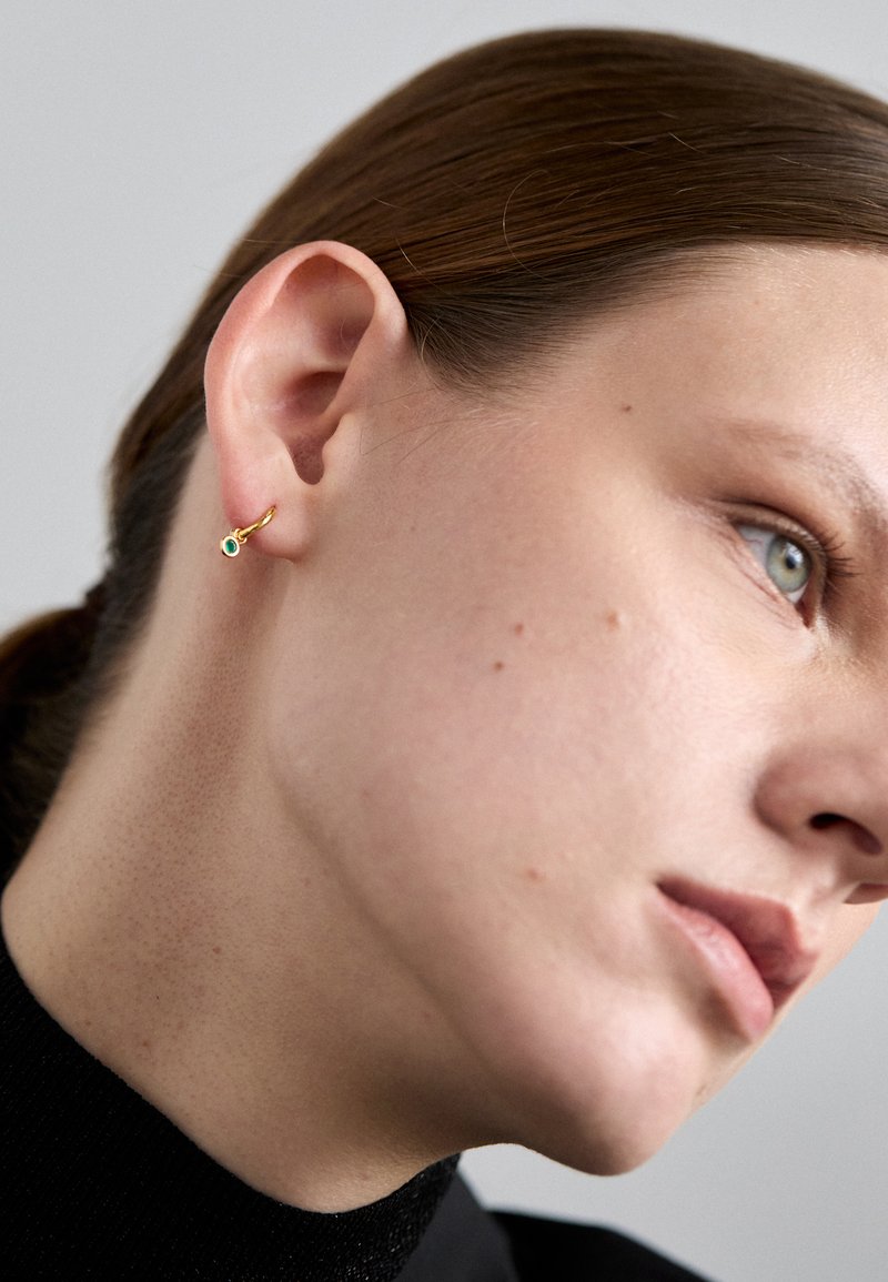 Brinco de ouro em forma de argola com um pequeno detalhe em turquesa. Close-up na orelha mostra uma textura de pele suave e uma parte superior escura.