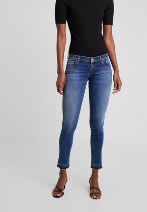 Femme portant un jean skinny bleu taille haute avec des ourlets effilochés, un haut noir à manches courtes et des sandales à talons noires à lanières, debout sur un fond gris.