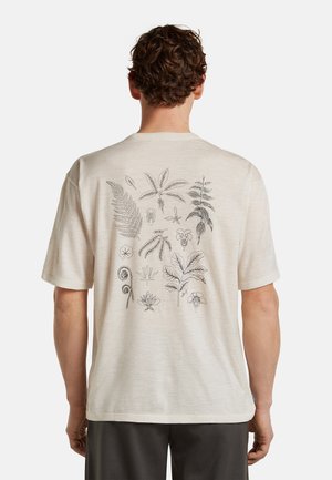Mężczyzna w beżowej koszulce z krótkim rękawem, na plecach czarne botaniczne szkice różnych liści i kwiatów.