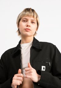 Jachetă din catifea neagră cu guler, buzunar frontal cu fermoar și patch cu logo alb. Purtați-o peste un tricou gri deschis cu guler rotund.