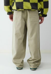 Brede beige broek van zwaar materiaal, met twee achterzakken en een iets taps toelopend ontwerp. Draagt met een gemusterde top.