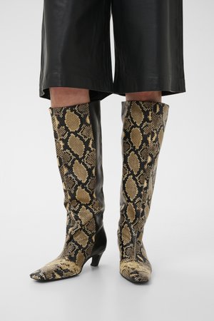 Kotníkové boty s hadím potiskem v béžové a černé barvě, nízkým podpatkem, nošené s černými koženými kalhotami nad kotníky.