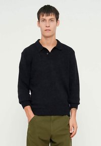 Musta neulottu neulepusero, jossa on kauluksellinen pääntie, 3/4-hihat ja teksturoitu viimeistely. Malli yhdistää sen vihreiden housujen kanssa.