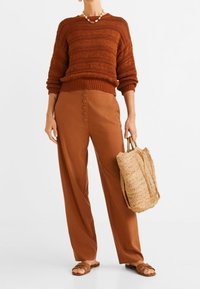 Sweter w kolorze rdzy, dziergany z fakturą, zestawiony z brązowymi, szerokimi spodniami z wysokim stanem i detalem guzikowym. Pleciona torba na ramię i sandały.