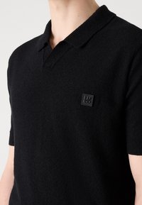 Zwarte polo shirt van textuurstof, met een korte kraag en korte mouwen. Het heeft een klein zwart leren logo op de borst.