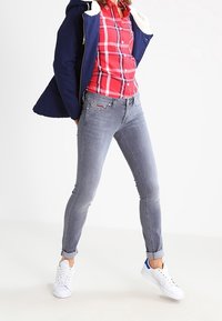 Jeans slim fit grigi con cinque tasche e passanti per cintura, abbinati a una camicia a quadri rossa sotto una giacca blu e sneakers bianche.