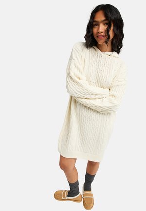 Young woman wearing a white cable knit sweater dress, gray socks, and tan shearling-lined slip-on shoes, standing with arms crossed.