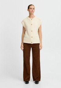 Cremfarbener, kurzärmeliger Strickcardigan mit gerippter Textur und fünf goldenen Knöpfen auf der Vorderseite, kombiniert mit braunen Cordhosens mit weitem Bein.