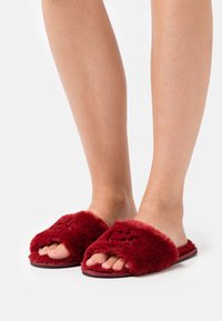 Chaussons glissants rouges et duveteux avec un design ouvert à l'avant, présentant une texture douce et une semelle épaisse. Rehaussés d'un matériau imitant la fourrure.