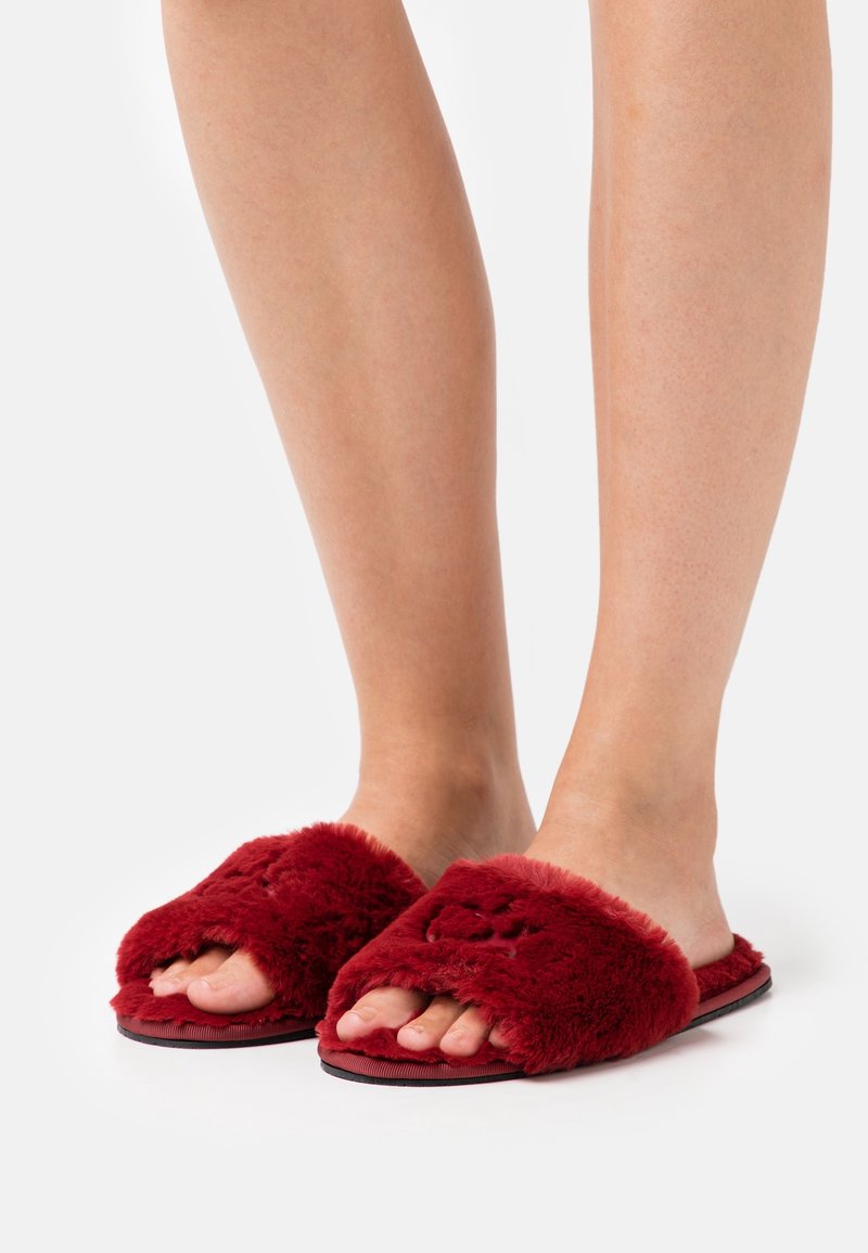 Chaussons glissants rouges et duveteux avec un design ouvert à l'avant, présentant une texture douce et une semelle épaisse. Rehaussés d'un matériau imitant la fourrure.